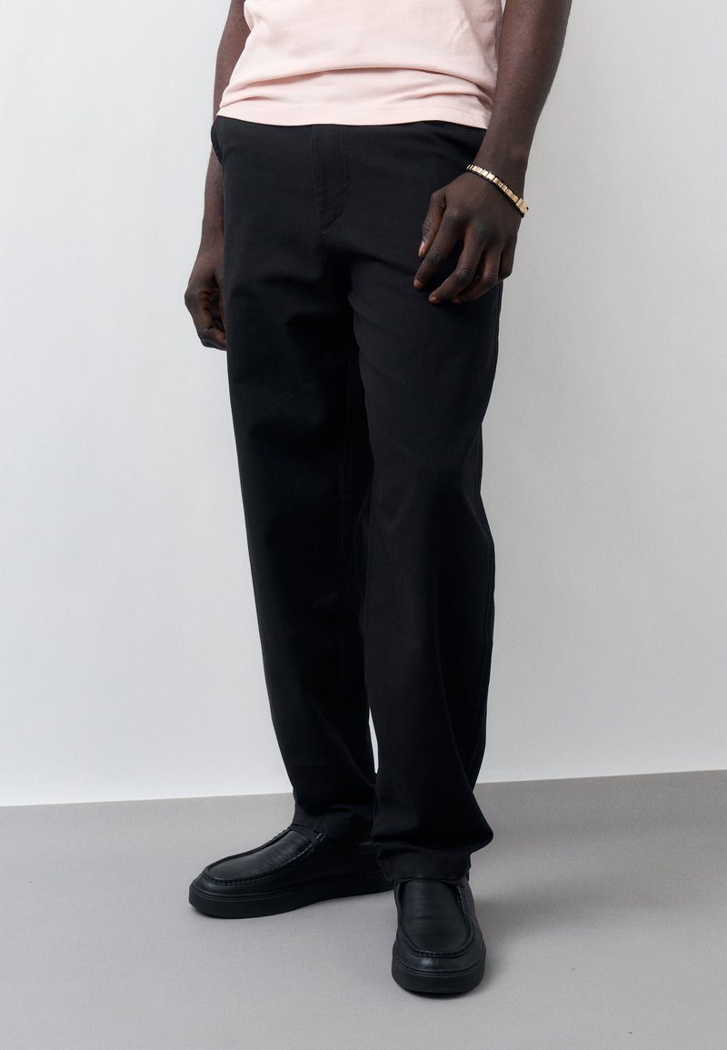 Άντρας που φοράει μαύρο παντελόνι ίσιας γραμμής, μαύρα παπούτσια χωρίς κορδόνια, ανοιχτό ροζ πουκάμισο και χρυσό βραχιόλι, στέκεται μπροστά σε ένα απλό φωτεινό φόντο.