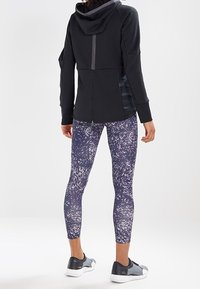 Veste noire texturée à fermeture éclair avec une capuche, associée à un legging bleu marine orné d'un imprimé abstrait blanc, et des chaussures de sport grises.