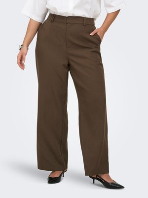 Mujer que lleva pantalones marrones de cintura alta y pernera ancha con trabillas, combinados con tacones negros puntiagudos y una blusa blanca de manga corta.
