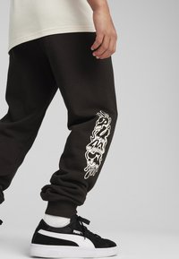 Černé joggingové kalhoty s elastickými manžetami, s bílým grafickým designem na straně. Nosí se s černými teniskami a bílými ponožkami.