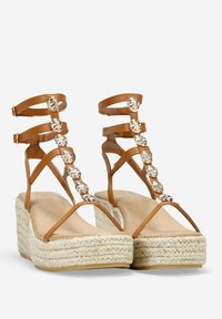 Sandalias de cuña de esparto con tiras marrones y suela de yute trenzado, que presentan adornos metálicos florales en la parte delantera y correas de tobillo ajustables.