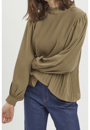 Kobieta w oliwkowej plisowanej bluzce z długim rękawem i wysokim kołnierzem oraz niebieskich dżinsach, pozująca z założonymi ramionami.