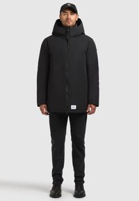 Svart vadderad jacka med hög krage och justerbar huva, frontdragkedja och sidofickor, kombinerad med svarta jeans och svarta stövlar.