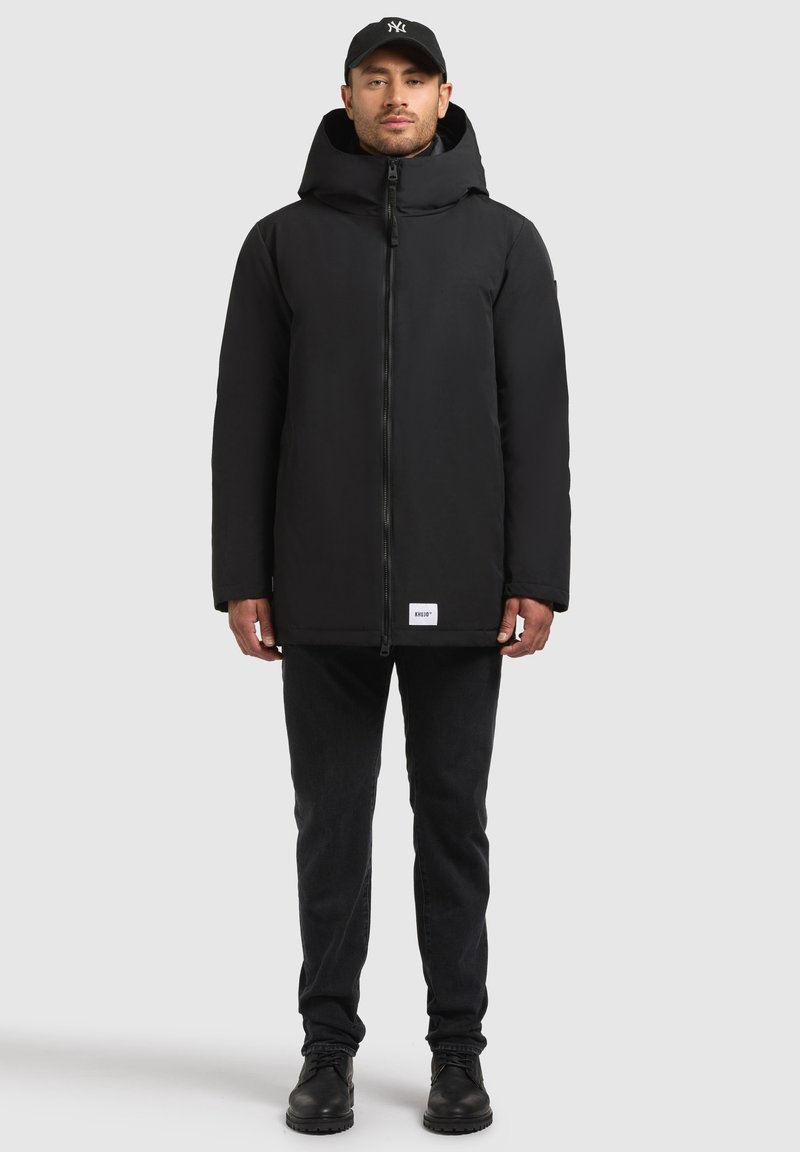 Svart vadderad jacka med hög krage och justerbar huva, frontdragkedja och sidofickor, kombinerad med svarta jeans och svarta stövlar.