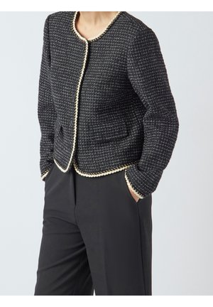 Kvinde iført sort tweedjakke med hvid kant og sorte bukser med høj talje, hænderne i lommerne, stående foran en ensfarvet baggrund.