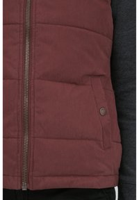 Gilet matelassé bordeaux avec fermeture éclair et poche à bouton-pression. Tissu texturé avec une finition mate, associé à une chemise à manches longues sombre.