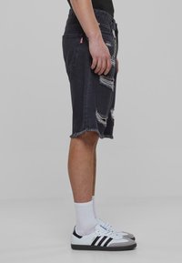 Pantalones cortos de mezclilla negra con detalles desgastados, bordes deshilachados y un diseño de tiro medio. Combinados con zapatillas blancas y calcetines blancos hasta la altura del tobillo.