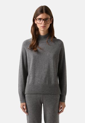 Femme portant de grandes lunettes, un pull à col montant en côte gris et un pantalon gris assorti, se tenant devant un fond clair.