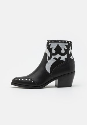 Bottine en cuir noir avec motifs décoratifs argentés et noirs, bordure cloutée, bout pointu et talon bloc sur fond blanc.