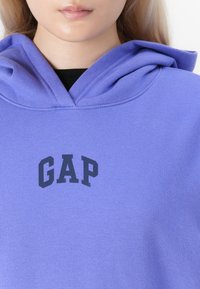 Personne portant un sweat à capuche violet avec le logo "GAP" en bleu marine, montrant la partie inférieure de son visage et son cou sur fond blanc.