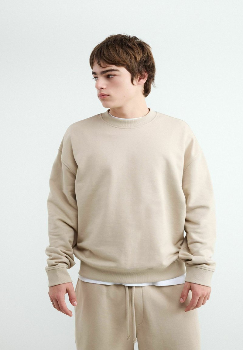 Jeune homme aux cheveux bruns portant un sweat beige et un pantalon de jogging assorti, regardant vers sa gauche sur un fond uni clair.