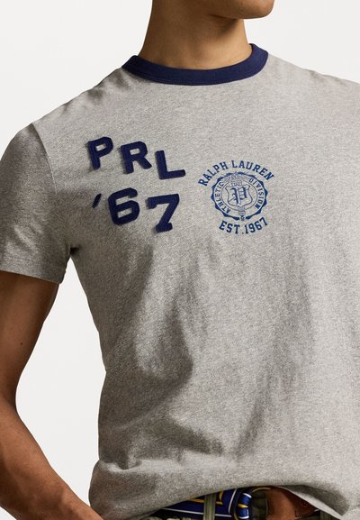 Pelēkas kokvilnas T-kreklu ar tumši zilu ribotu apkakli, kuram ir tumši zila izšūta uzraksts "PRL '67" un apaļš logo prints uz krūšu daļas.