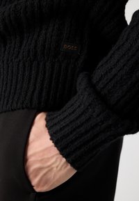 Μαύρο πλεκτό πουλόβερ με ριγωτή υφή, με ένα διακριτικό λογότυπο. Επικαλυμμένο πάνω από μαύρο παντελόνι με το χέρι στη τσέπη.