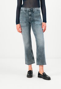 Personne portant un jean gris taille haute et court avec des poches avant en patch, un haut noir texturé à manches longues et des mocassins noirs brillants sur un sol blanc.