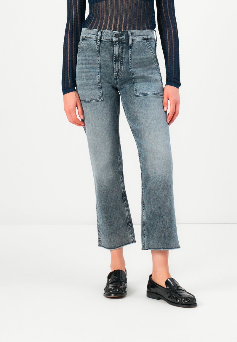 Personne portant un jean gris taille haute et court avec des poches avant en patch, un haut noir texturé à manches longues et des mocassins noirs brillants sur un sol blanc.