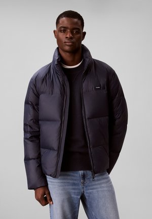 Homme portant une doudoune foncée sur un pull noir et un jean bleu clair, debout devant un fond gris uni.