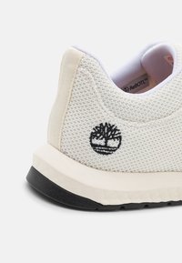 Vit mesh-sneaker med texturerad yta, med en svart broderad logotyp och en mjuk, krämfärgad mellansula samt svart yttersula.