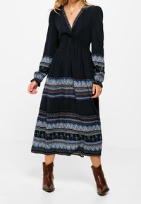 Pitkä musta mekko, jossa on V-pääntie, kierretty vartalo ja pitkät hihat. Sisältää sinisistä ja vihreistä koostuvan kuviollisen helman, jossa on koristeellisia yksityiskohtia.