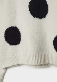 Pull en maille blanche avec de grands pois noirs. Le tissu semble doux avec un détail de bord-côte à l'ourlet, offrant une texture confortable.