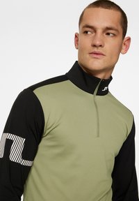 Långärmad träningskavaj med dragkedja i kragen, olivgrön texturerad framsida och svarta ärmar med vitrandig logodetalj.