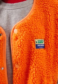 Erksav oranžist fliisvest, mis on kinnitatud trukkidega ja kantud halli dressipluusi peal, mille rinnal on silt "La Maison AMV Super Vintage".