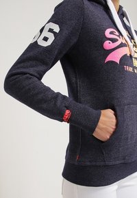 Sudadera marinera con cordón blanco, gran número "96" blanco en la manga y etiqueta de la marca roja en el puño. Tejido suave y texturizado.