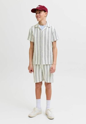 Tiener met een bordeauxrode pet, een wit overhemd met knopen en een korte broek met verticale zwarte strepen, witte sokken en sneakers, staand en glimlachend.