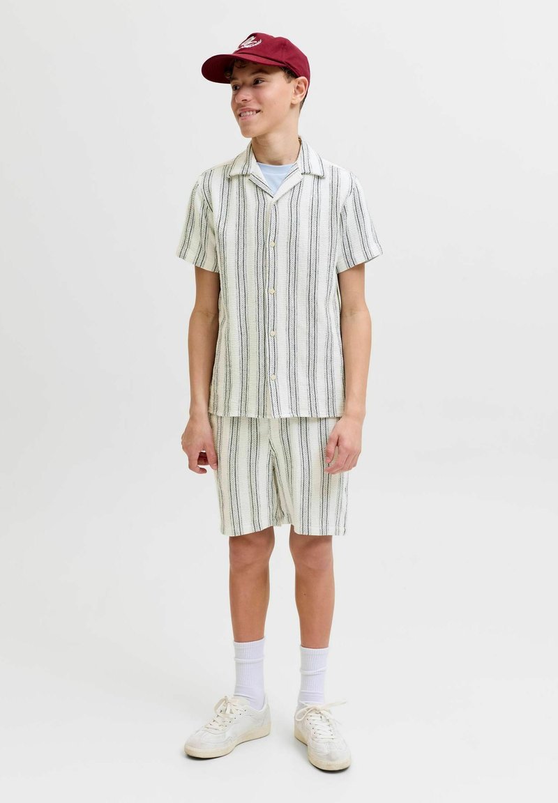 Adolescent portant une casquette bordeaux, une chemise blanche à boutons et un short à rayures verticales noires, chaussettes blanches et baskets, debout et souriant.