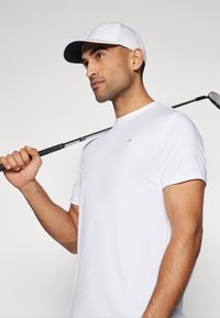 Camisa de golfe branca de manga curta com decote redondo, textura suave, combinada com uma chapa branca e azul-marinho. Segurando um taco de golfe sobre o ombro.
