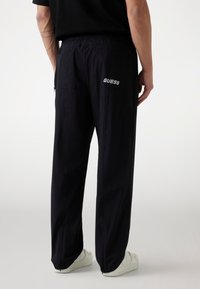 Black jogger pants with an elastic waistband, zippered pockets, and logo detail on the back. Smooth fabric with ankle zippers.
