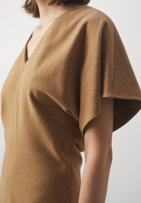 Femme portant une robe courte marron chameau à manches courtes, avec un col en V, un tissu texturé et un design à manches supérieures amples, sur un fond uni.