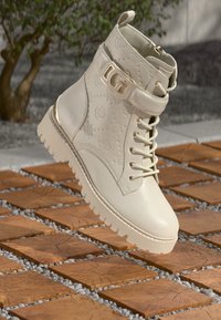 Botín de cuero blanco hasta el tobillo con cierre de cordones en la parte delantera, suela gruesa, detalles en dorado y un patrón floral en relieve.