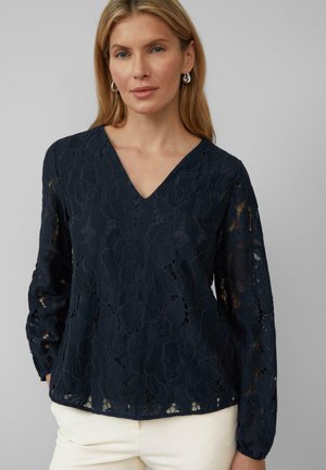Blouse bleu marine avec un col en V, un motif de dentelle florale complexe, des manches longues avec des poignets élastiques, et un tissu léger et texturé.