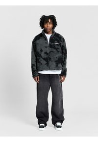 Giacca in pile zip-up nera con motivo mimetico grigio; indossata sopra una maglietta bianca. Pantaloni jeans neri larghi e sneakers bianche e nere.