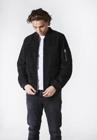 Svart mocka bomberjacka med ribbade ärmslut och nederkant. Har en ficka med dragkedja på vänster ärm. Bärs över en vit T-shirt och mörka jeans.