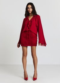 Blouse rouge avec un devant froncé et de longues manches évasées, associée à une mini jupe rouge texturée ornée de motifs de roses. Talons aiguilles rouges.