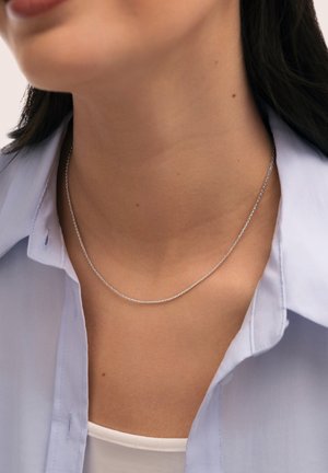 Primer plano de una mujer que lleva un collar de cadena fina de plata, una camisa azul clara con el cuello abierto y una camiseta interior blanca.