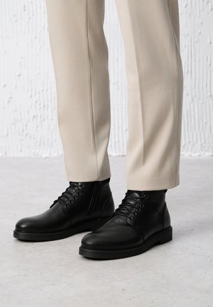 Czarne skórzane botki za kostkę o teksturowanym wykończeniu, okrągłym nosku, sznurowanej budowie i kontrastowym przeszyciu, zestawione z jasnymi dopasowanymi spodniami.
