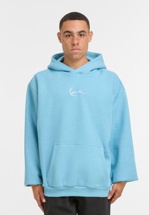 Uomo con capelli corti e lentiggini che indossa un felpa blu chiaro con tasca frontale e testo bianco sulla maglia, in piedi davanti a uno sfondo bianco.
