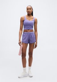 Γυναίκα στέκεται κρατώντας ροζ λάστιχο αντίστασης, φοράει μωβ αμάνικο αθλητικό τοπ και σορτς, λευκές κάλτσες και λευκά αθλητικά παπούτσια σε απλό φόντο.