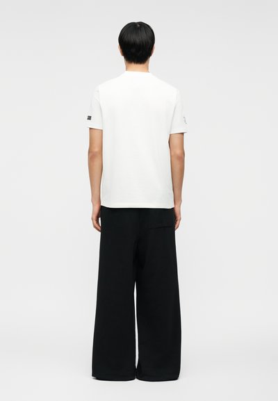 Άτομο με κοντά μαύρα μαλλιά, φορώντας απλό λευκό κοντομάνικο μπλουζάκι και φαρδιά μαύρη παντελόνα, όρθιο με την πλάτη προς τα εμπρός.