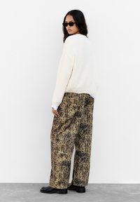 Krém színű kötött pulóver, laza bézs nadrág leopárdmintával és fekete bokacsizma. A modellen sötét napszemüveg van, háttal áll.