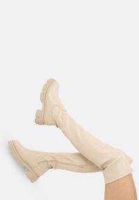Botas beiges hasta la rodilla hechas de material suave, con suela gruesa y detalle de cremallera lateral. El diseño es ajustado, abrazando la pierna.