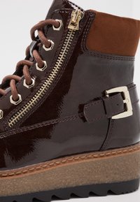 Hnědé lakované kožené kotníkové boty s kovovým zipem, hnědými tkaničkami, dekorativní přezkou a texturovanou, světle zbarvenou klínovou podrážkou.