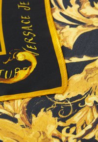 Silkkihuivi, jossa on musta tausta ja rohkea kultainen barokkikuosi, jota ympäröi keltainen ommeltu reuna ja Versacen logo -yksityiskohta.