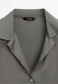 Col de chemise gris foncé avec revers ouverts et un seul bouton visible, montrant des bords de tissu légèrement effilochés près de la patte de boutonnage.
