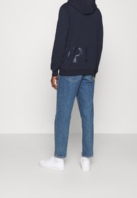 Man i marinblå hoodie med "POP!" tryck, blå jeans, vita strumpor och vita sneakers, står framför en enkel ljusgrå bakgrund.