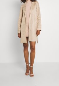 Cappotto lungo in suede beige dalla texture morbida, con rever a punta e maniche lunghe, abbinato a un abito a coste rosa e sandali beige con cinturini.