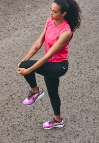 Žena se protahuje při stání na asfaltu, oblečená v růžovém bezrukávovém topu, černých legínách a růžových běžeckých botách s černými tkaničkami.
