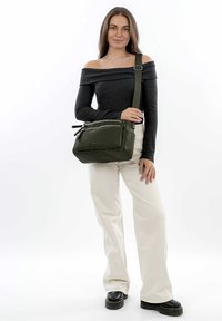 Borsa a tracolla verde in tessuto testurizzato, con tasca frontale con zip e tracolla regolabile, posizionata su uno sfondo neutro.